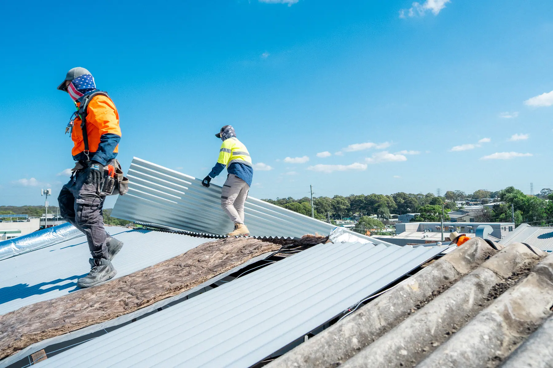 Two roofers carrying a new metal roof sheet across the old fibre-cement roof