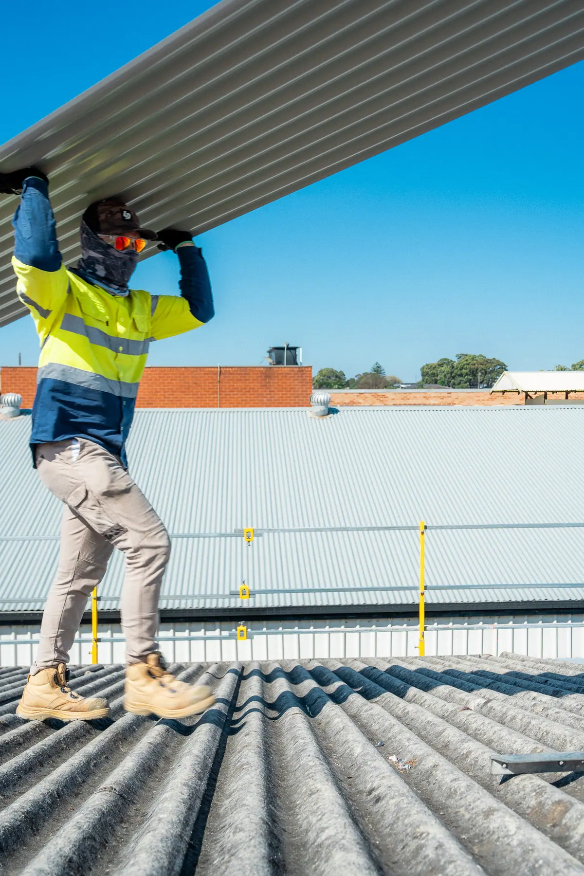 Roofer in yellow hi-vis carrying a metal sheet overhead across the old fibre-cement roof