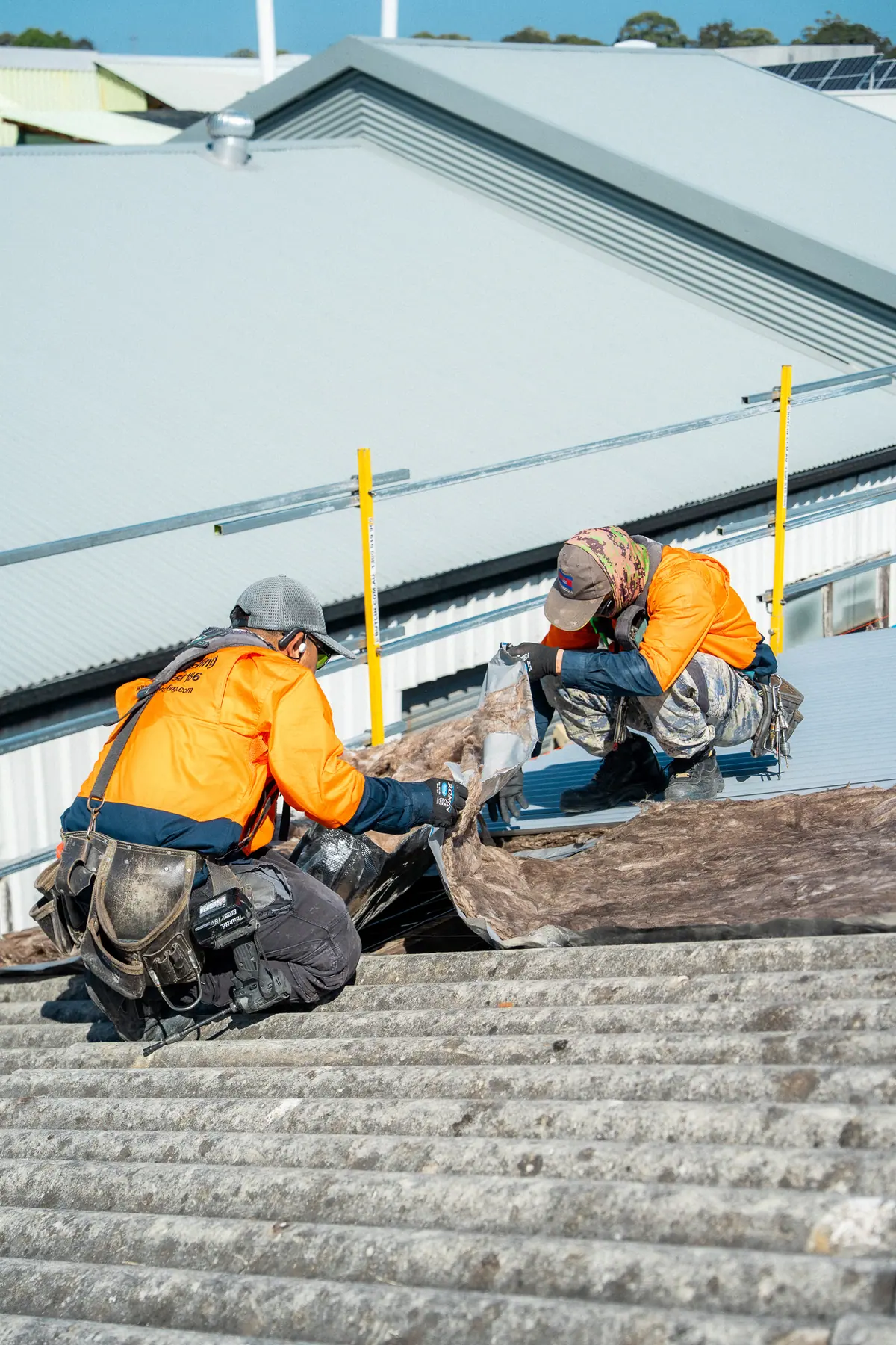 Two roofers kneeling on the old fibre-cement roof beside the edge protection
