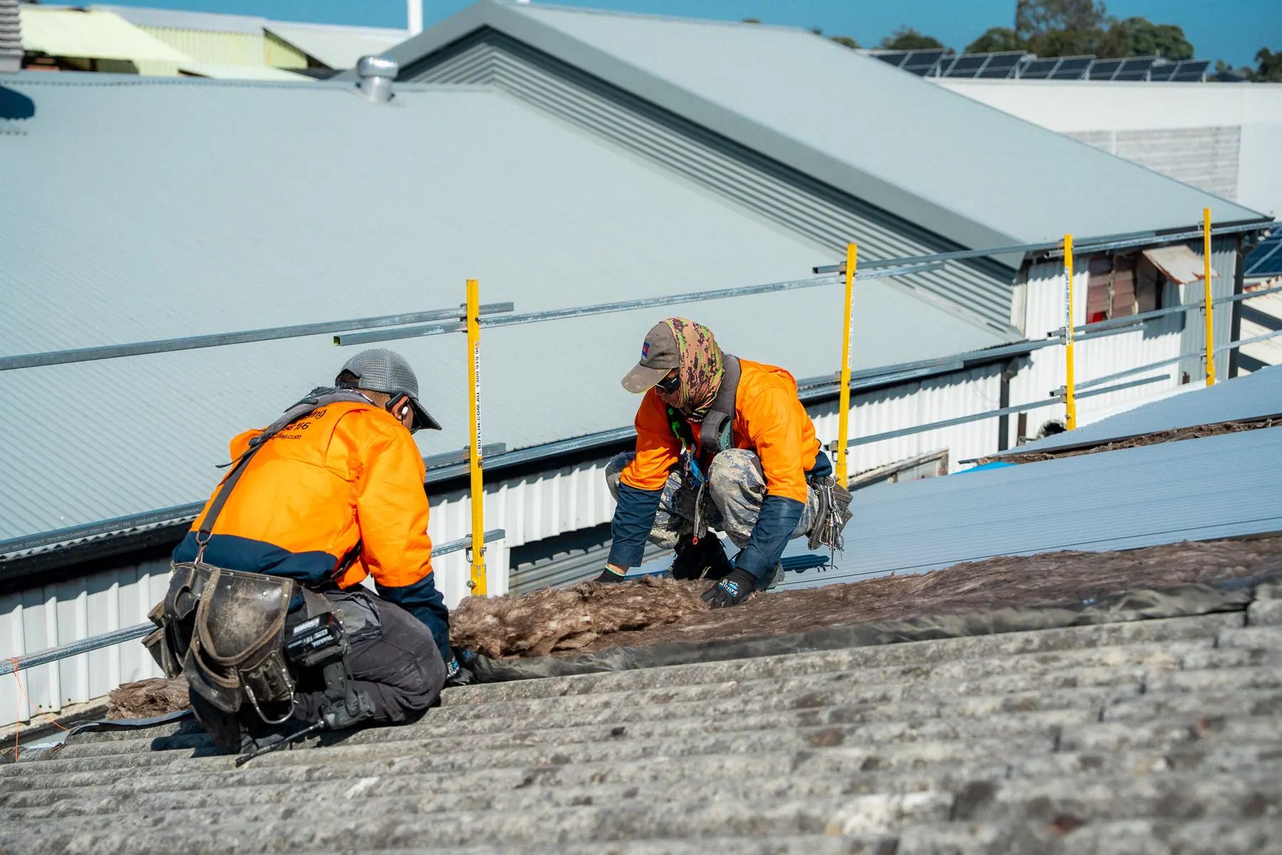 Two roofers kneeling on the old fibre-cement roof as stripping begins