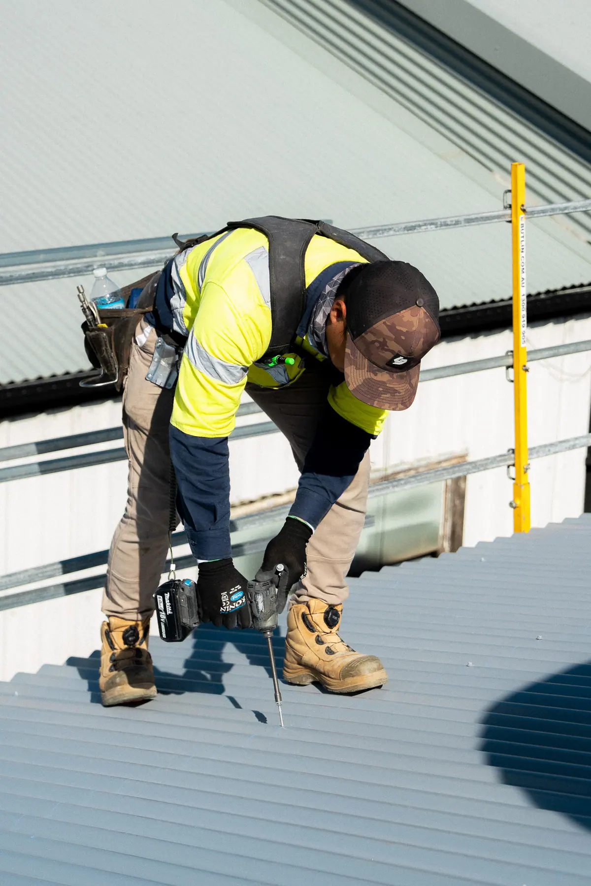 Roofer in yellow hi-vis screwing down new sheets near the edge rails