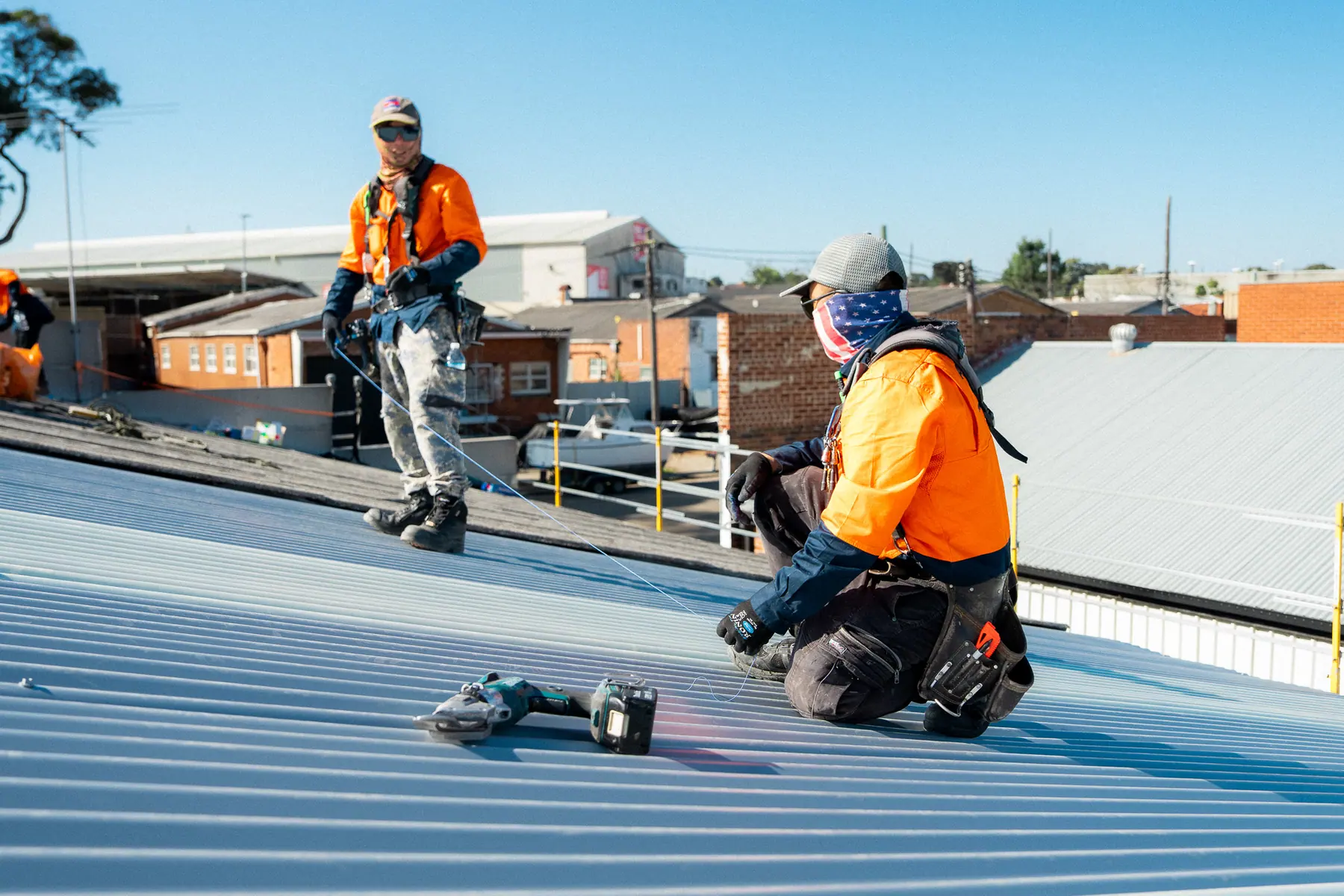 Roofer kneeling with a drill while a crewmate checks the sheet line